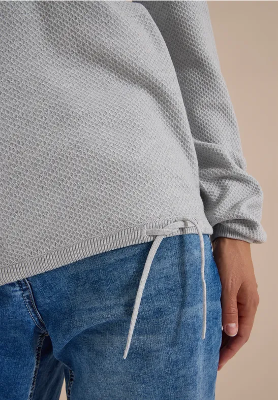 Gros plan d’un pull gaufré gris à cordon de serrage à la base, associé à un jean bleu.