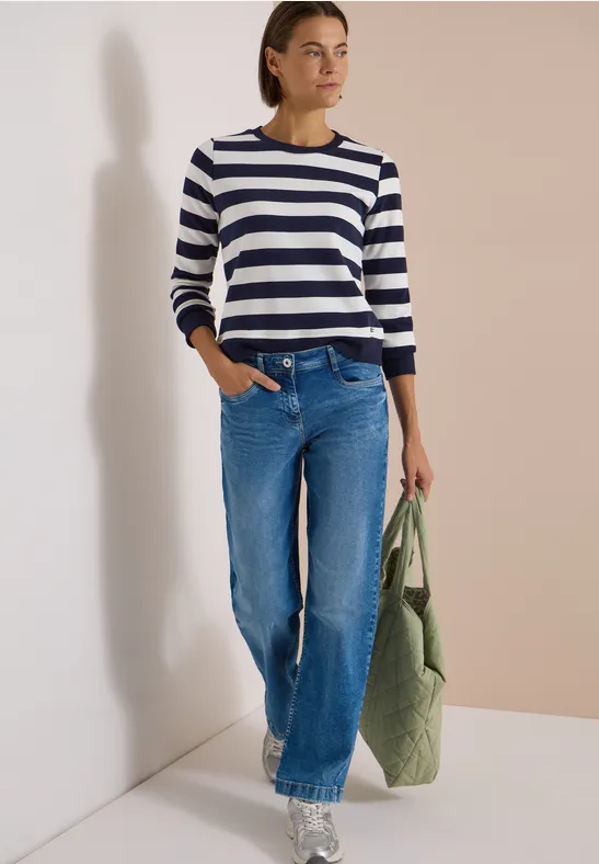 Frau trägt gestreiften Pullover, weite Jeans und hält eine grüne, gesteppte Stofftasche vor neutralem Hintergrund.