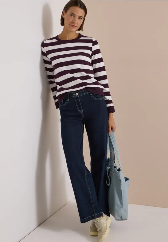 Frau lehnt an Wand, trägt gestreiften Pullover, weite Jeans und hält eine hellblaue Tasche.