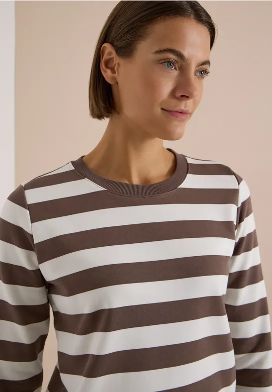 Frau trägt ein braun-weiß gestreiftes Langarmshirt, helles Studio-Setting, leicht lächelnd, kurze Haare.
