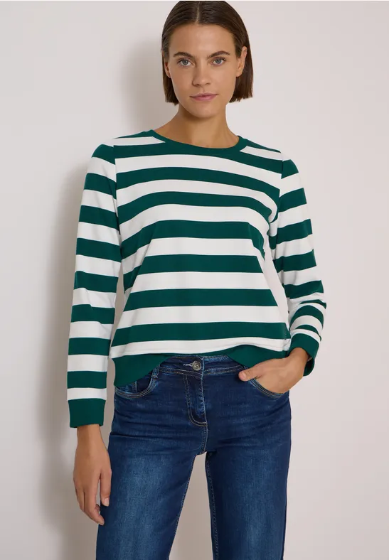 Frau in grün-weiß gestreiftem Pullover und blauer Jeans vor schlichtem Hintergrund.