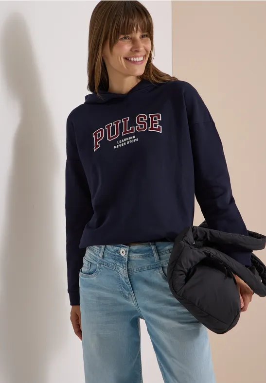 Femme souriante en sweat à capuche bleu marine « PULSE », jean clair et veste matelassée noire à la main.
