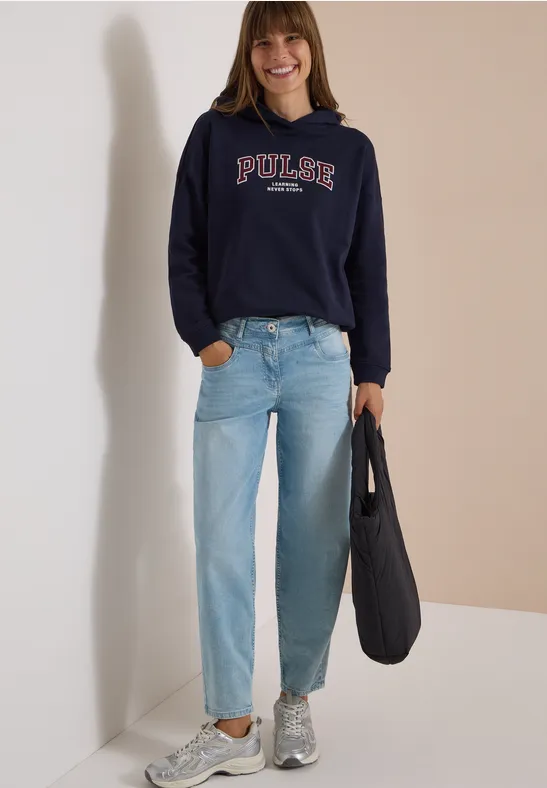 Une femme souriante porte un sweat bleu marine 'PULSE', un jean clair, des baskets grises et un sac noir.