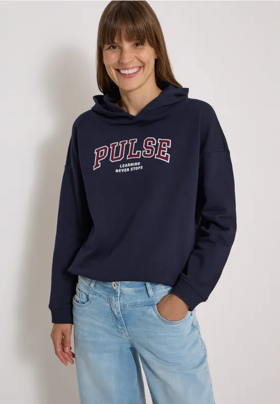 Femme en sweat à capuche bleu marine 'PULSE' et jean bleu, souriante sur fond clair.
