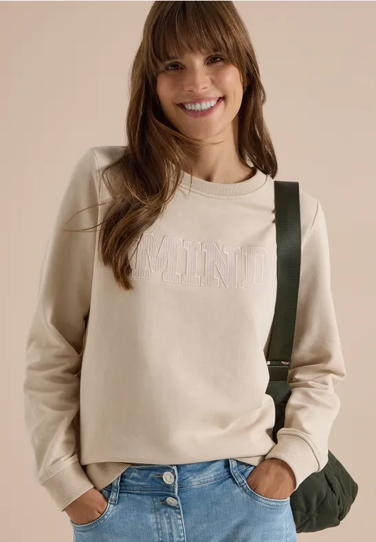 Femme souriante aux cheveux bruns portant un pull beige, un jean et un sac bandoulière.