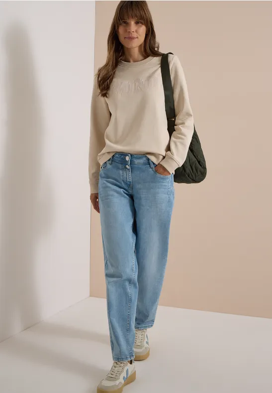 Femme en pull beige, jean bleu et baskets, portant un sac vert devant un mur beige clair.