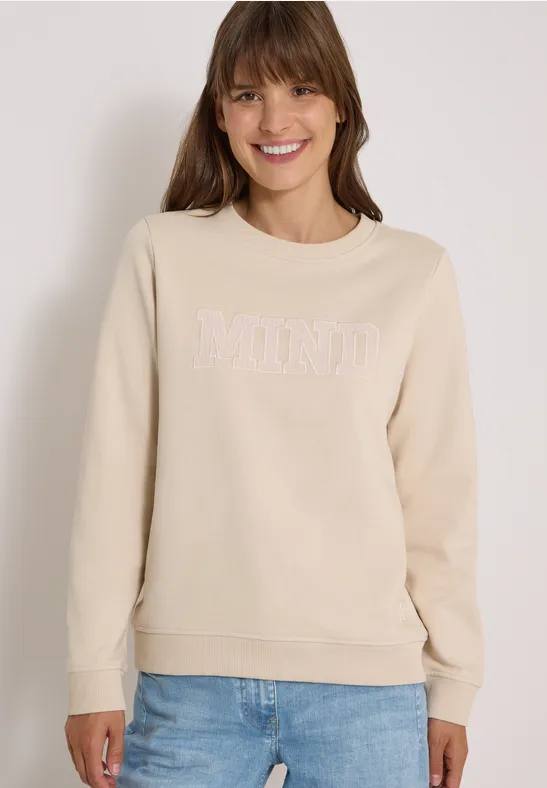 Femme en sweat-shirt beige 'MIND' et jean bleu, souriante sur fond clair.