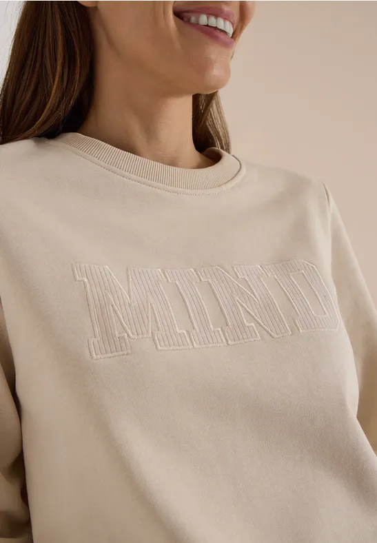 Femme de profil porte un sweat écru brodé «MIND» et sourit légèrement.
