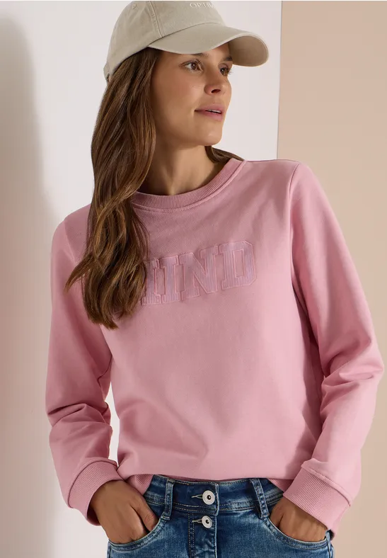 Femme porte un sweat rose, un jean bleu et une casquette beige.
