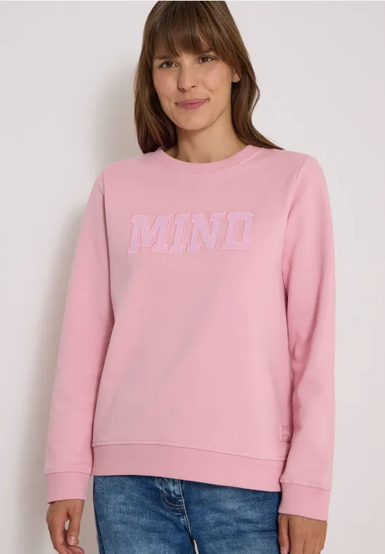 Femme portant un sweat-shirt rose avec l’inscription blanche "MIND", souriante.