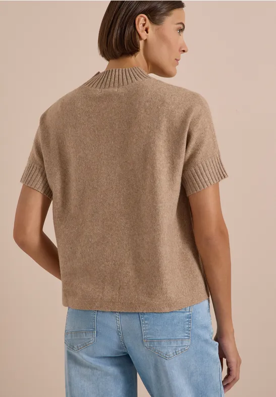 Femme vue de dos portant un pull court beige en maille à manches courtes et un jean bleu clair sur fond beige.