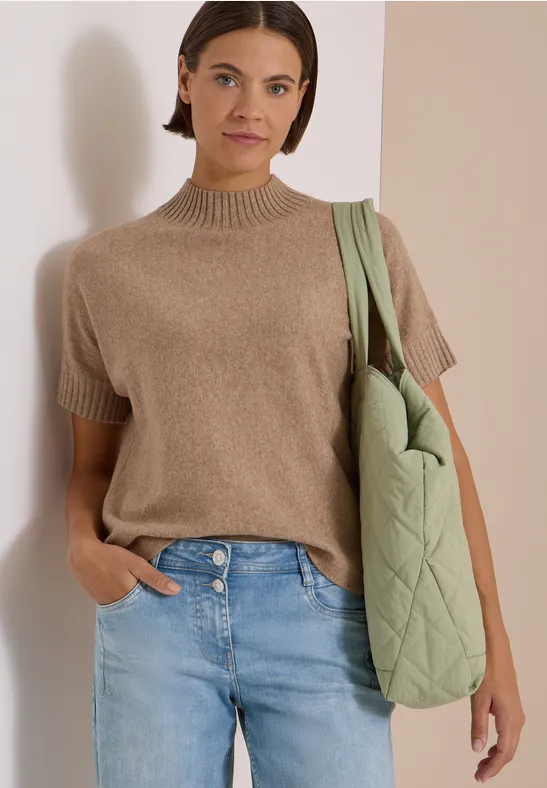 Femme en pull court beige, jean clair et grand sac matelassé vert, appuyée contre un mur.