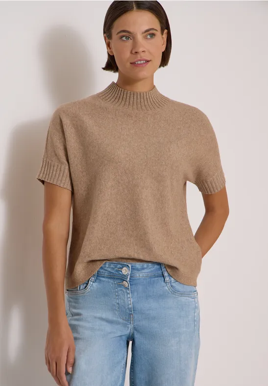 Une femme porte un pull en maille beige à manches courtes et un jean bleu devant un fond clair.