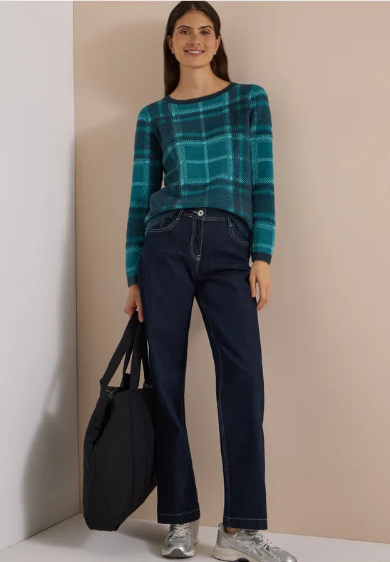 Frau in kariertem grünen Pullover, weiten dunkelblauen Jeans und großer Tasche.