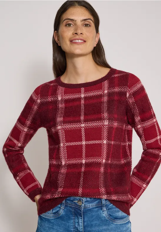 Frau mit rotem kariertem Pullover und Jeans, die lächelnd Hände in den Taschen zeigt.
