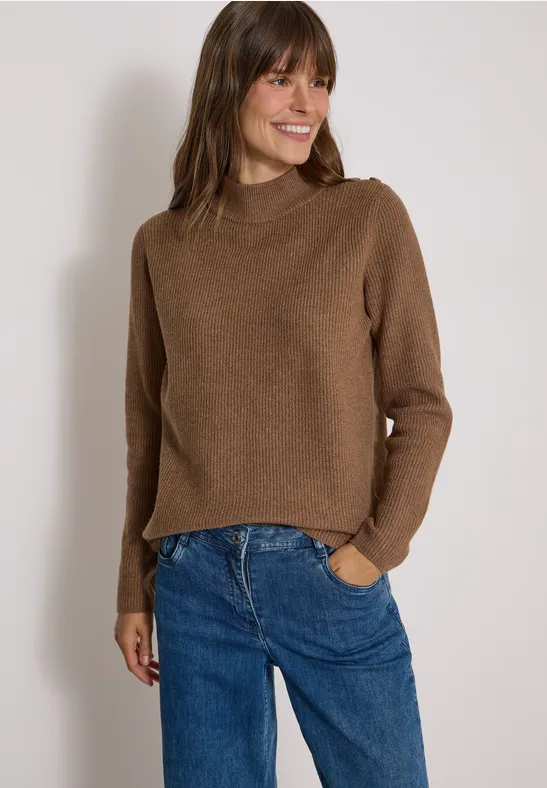 Une femme souriante porte un pull marron et un jean bleu sur fond clair.