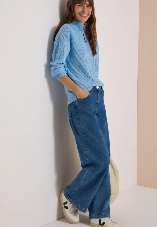 Femme adossée à un mur, pull bleu clair, jean large et baskets blanches, sourire décontracté.