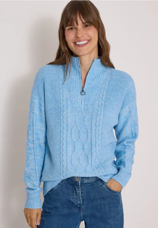 Femme en pull bleu à col zippé et jean, souriante, les mains dans les poches.