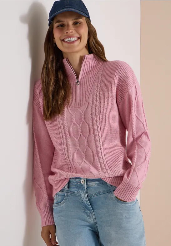 Une femme souriante portant un pull rose torsadé à col zippé, un jean et une casquette bleu marine.
