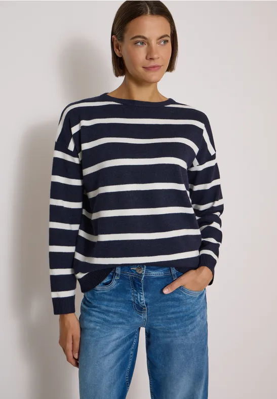 Frau trägt navyweiß gestreiften Pullover und blaue Jeans vor weißem Hintergrund.