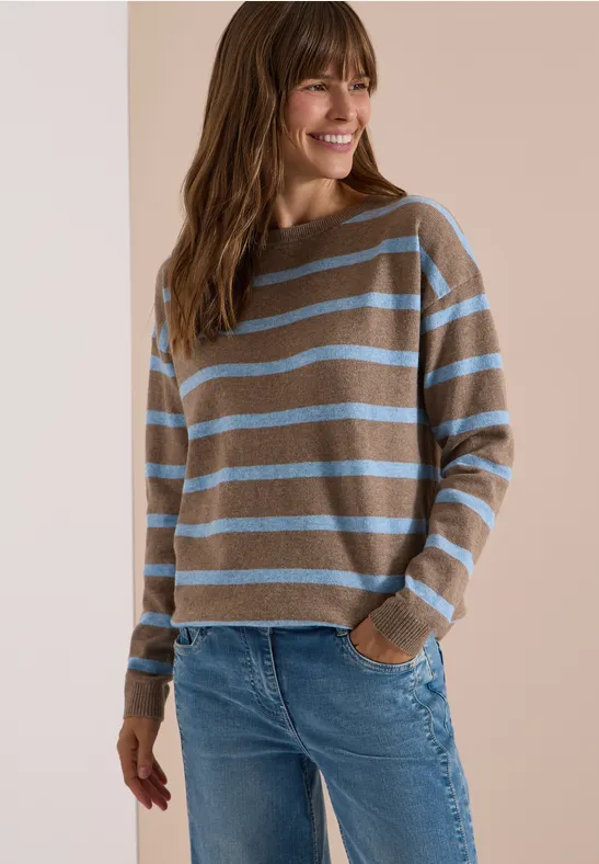 Eine lächelnde Frau trägt einen grauen Pullover mit hellblauen Streifen und blaue Jeans.