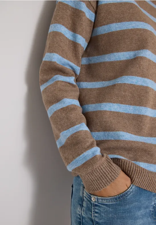 Person trägt braun-blauen gestreiften Pullover und Jeans, Hand in Hosentasche.