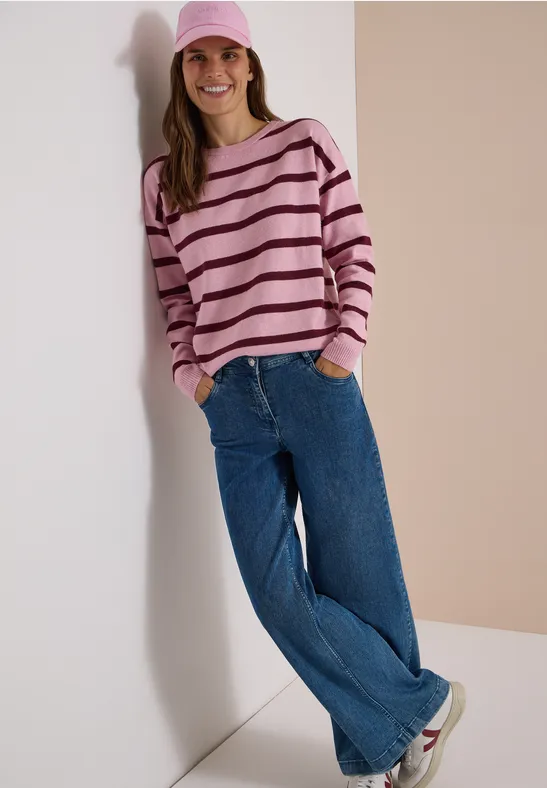 Femme souriante appuyée contre un mur, casquette rose, pull rayé, jean large et baskets blanches.