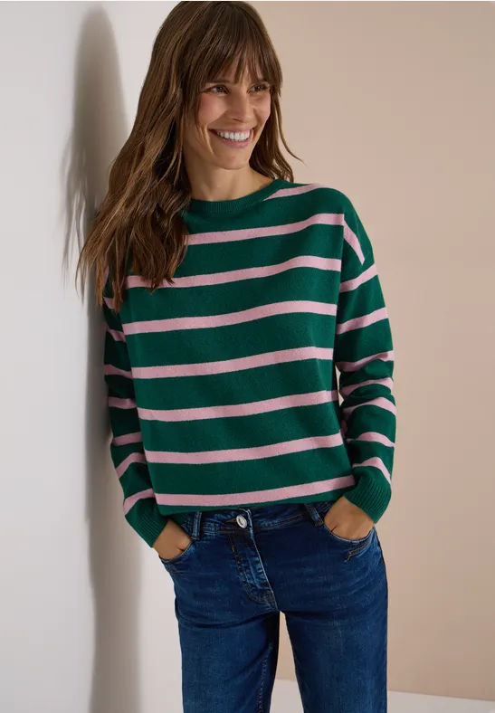 Femme souriante adossée à un mur, porte un pull rayé vert et rose et un jean bleu.