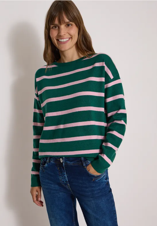Femme souriante en pull vert à rayures roses et jean.