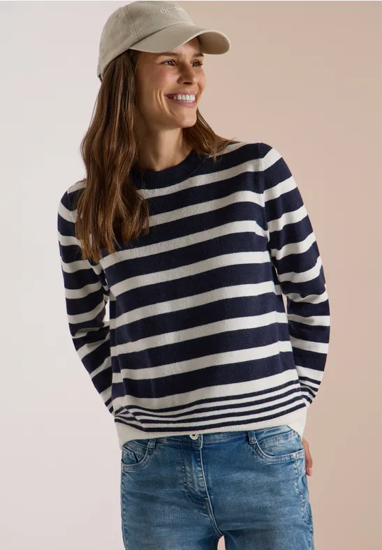 Une femme souriante porte un pull rayé, un jean et une casquette devant un fond neutre.