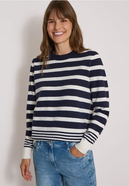 Une femme souriante porte un pull à rayures bleu marine et blanc avec un jean devant un mur blanc.