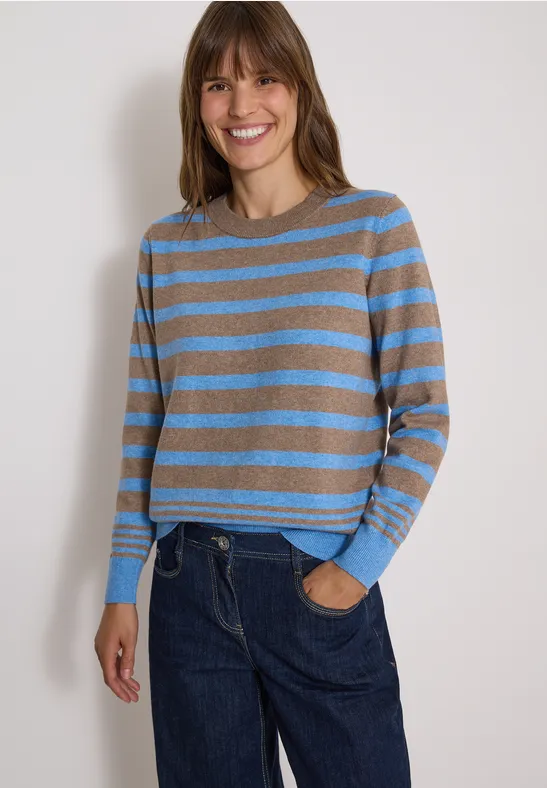 Une femme sourit en pull à rayures bleu et gris avec un jean.
