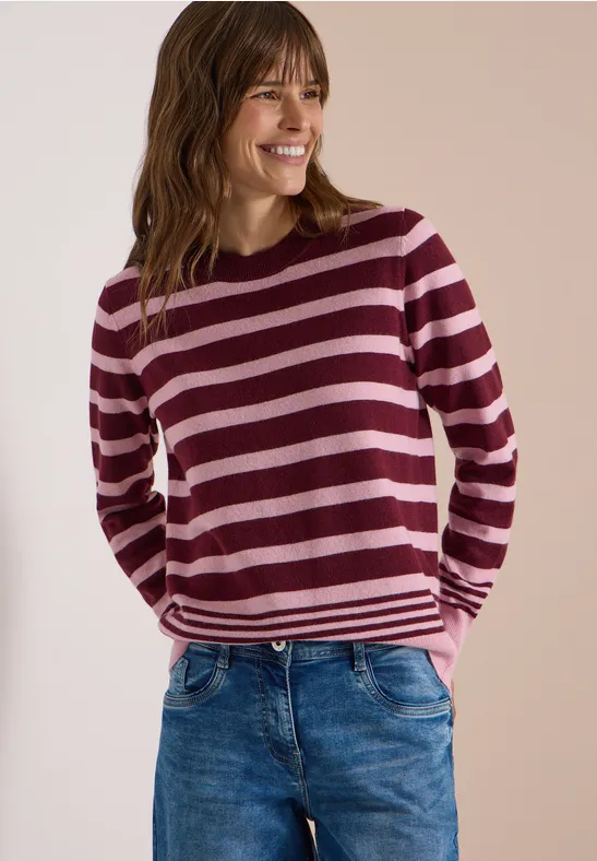 Femme souriante portant un pull rayé rose et bordeaux avec un jean, fond sobre neutre.