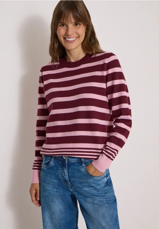 Une femme sourit devant un fond clair, portant un pull rayé rose et bordeaux avec un jean.
