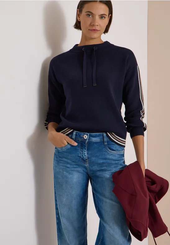 Eine Frau in blauem Pullover mit Kordelzug und Jeans hält eine bordeauxrote Jacke.