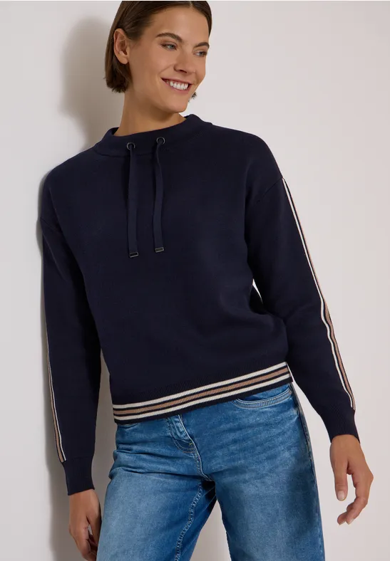 Frau in blauem Kapuzenpullover mit Streifen an Ärmeln und Bund, kombiniert mit Jeans, steht lächelnd.