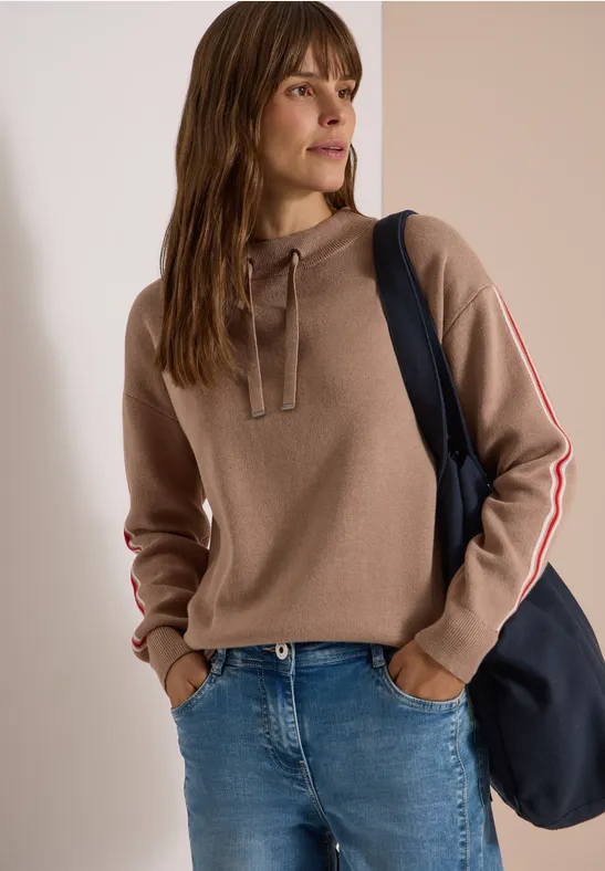 Frau in taupefarbenem Hoodie mit gestreiften Ärmeln, Jeans und dunkelblauer Tasche.
