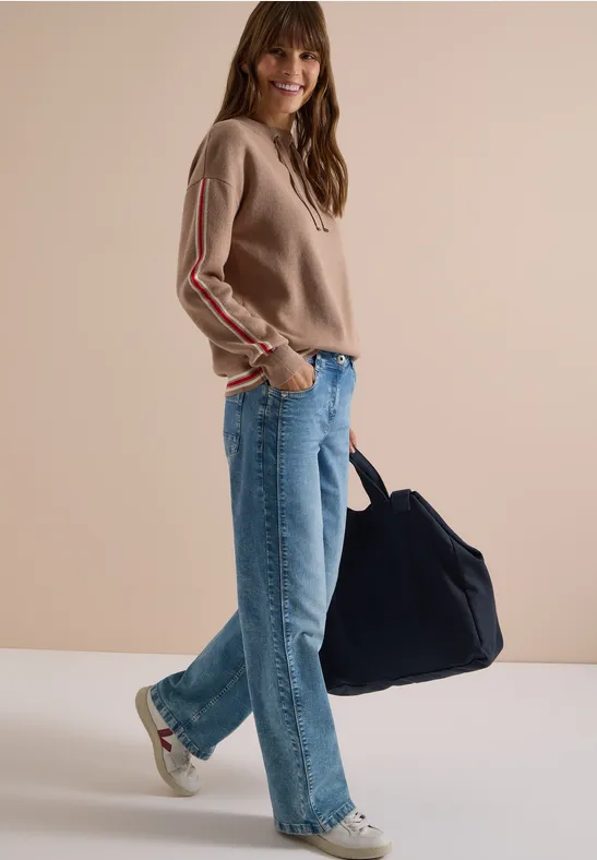Frau in beigem Pullover, blauen Jeans, Sneakern, trägt große blaue Tasche, vor schlichter Wand lächelnd.