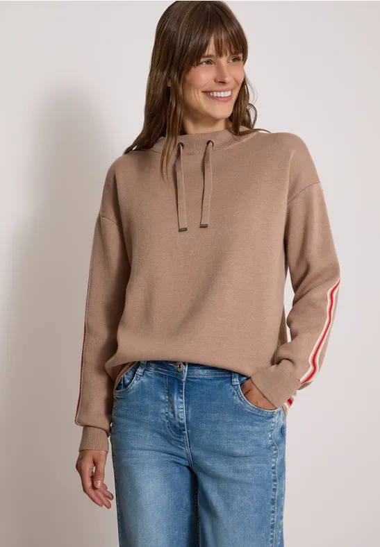 Frau in beigem Hoodie mit Streifen am Ärmel und blauen Jeans, lächelnd.