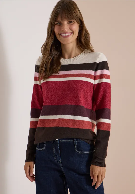 Une femme souriante porte un pull rayé rouge, rose et marron avec un jean, devant un fond clair.