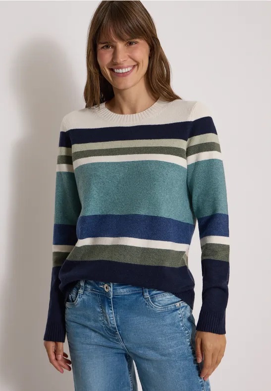 Femme souriante portant un pull à rayures bleues, vertes et blanches avec un jean, sur fond clair.