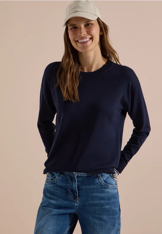 Femme souriante en casquette blanche, pull bleu marine et jean, posant de manière décontractée.