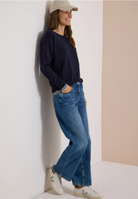 Une femme souriante en jean, pull bleu marine et casquette, adossée à un mur clair.