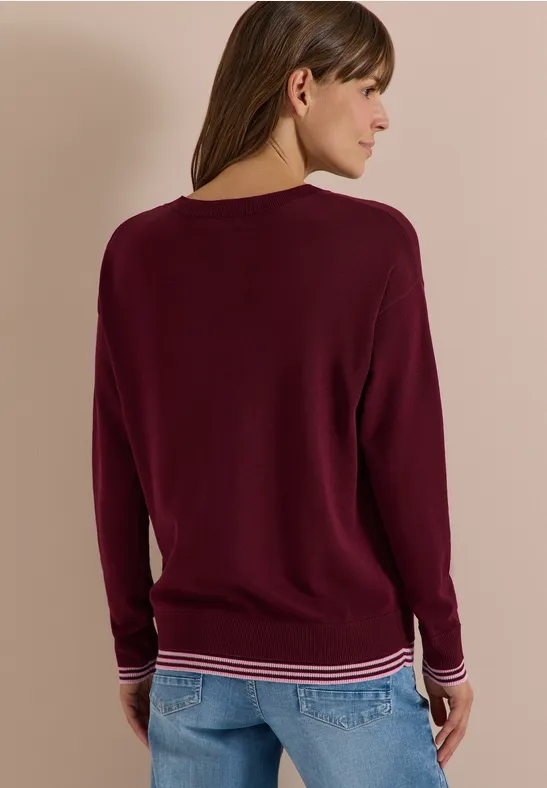 Frau von hinten in bordeauxrotem Pullover mit gestreiftem Bund und Jeans.