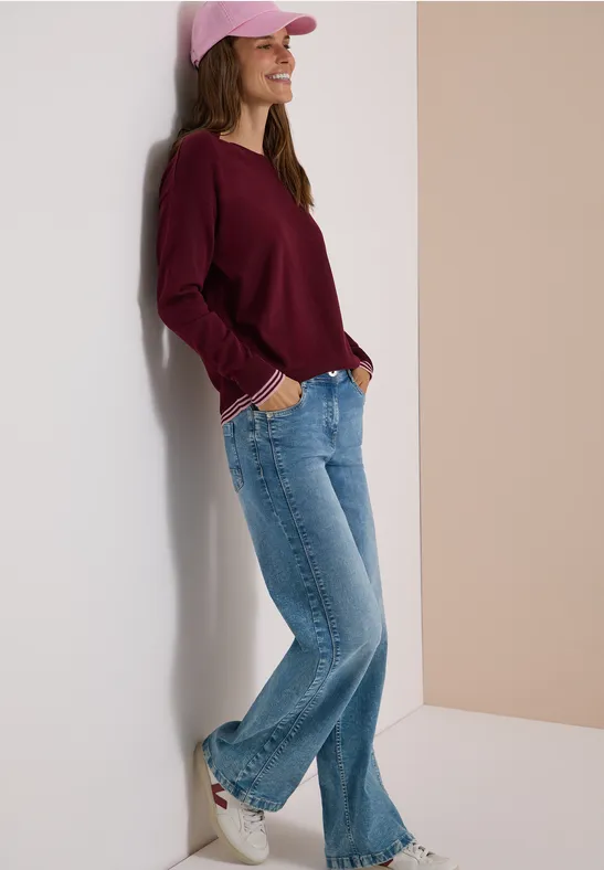 Eine lächelnde junge Frau in Jeans, burgunderfarbenem Pullover, rosa Cap und Sneakern lehnt entspannt an der Wand.
