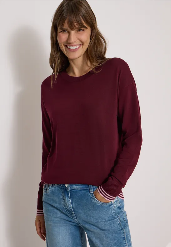 Lächelnde Frau in weinrotem Langarmshirt und Jeans vor hellem Hintergrund.