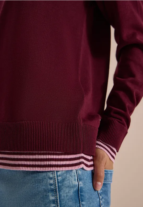 Roter Pullover mit gestreiftem Saum und Jeans, rechte Hand in der Hosentasche.