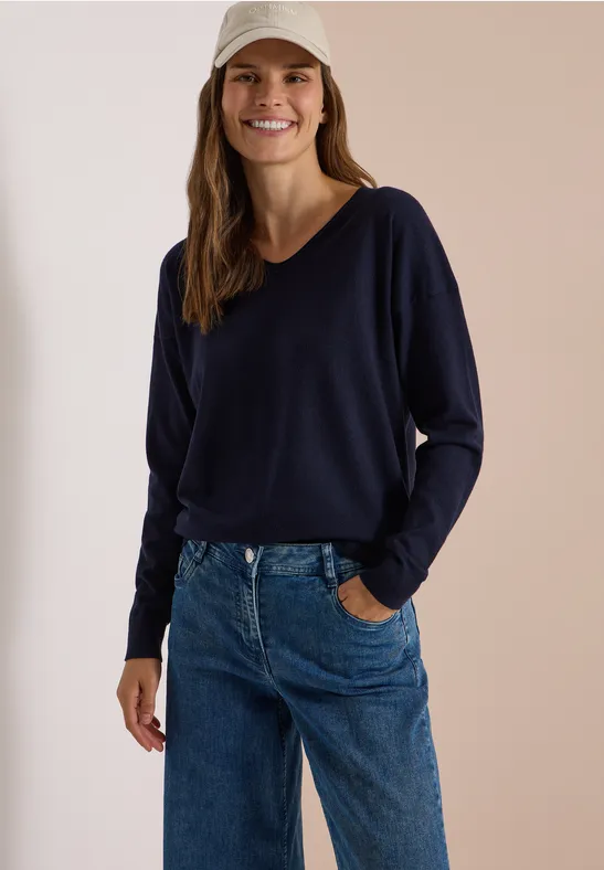 Femme souriante en pull bleu marine, jean et casquette beige.