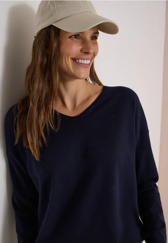 Femme souriante appuyée sur un mur, casquette beige et pull bleu foncé.