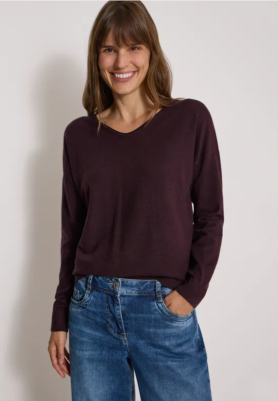Fröhliche Frau trägt bordeauxfarbenen Pullover und Jeans, posiert lächelnd vor neutralem Hintergrund.